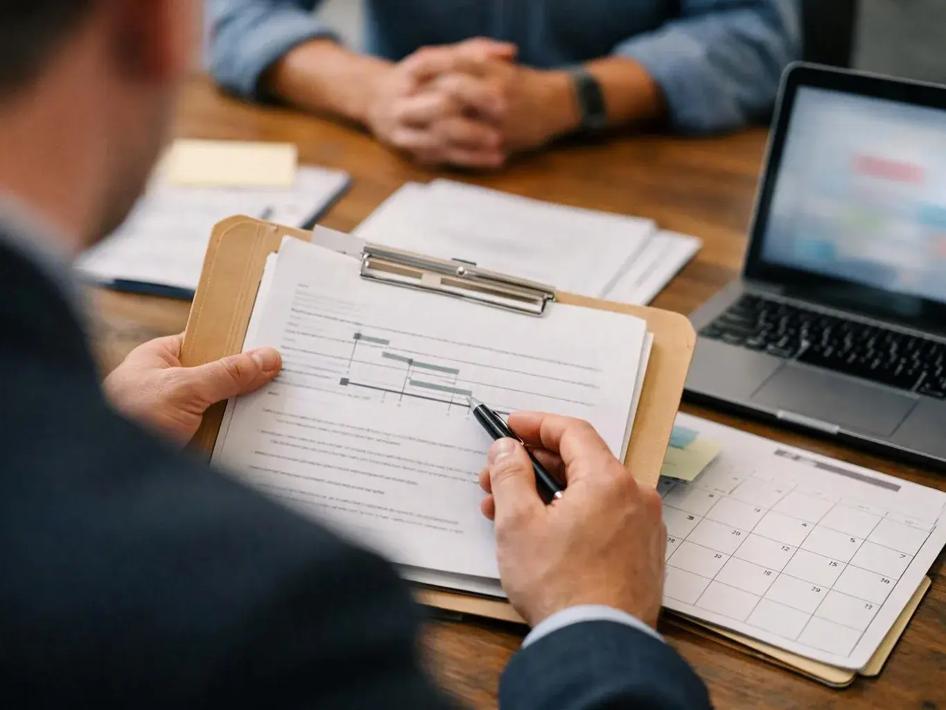 A person points at a printed chart on a clipboard during a meeting at a wooden table with a laptop nearby