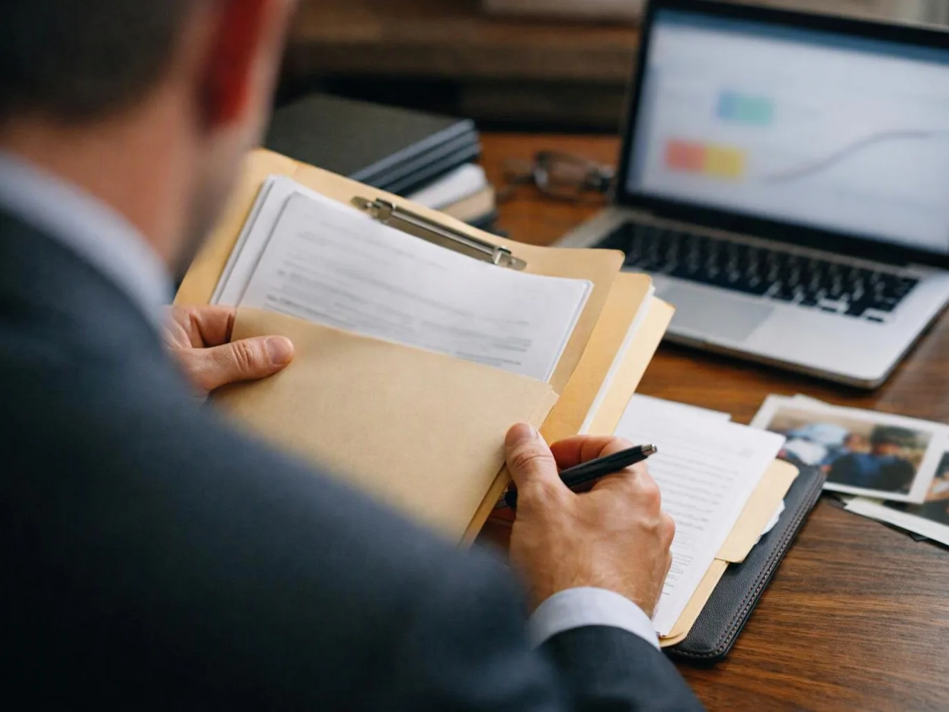 A person in a suit reviews papers on a clipboard beside an open laptop at a wooden desk
