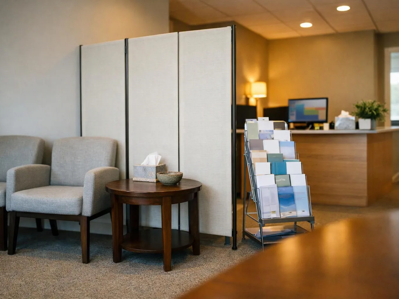 A quiet office waiting area shows two gray chairs, a round table, brochure rack, and reception desk behind a divider