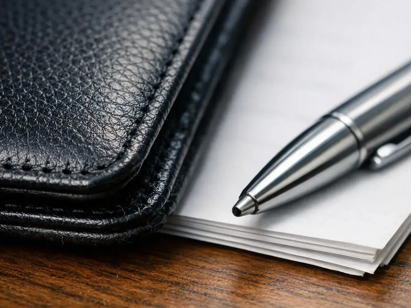 A silver pen rests beside a black leather wallet on a stack of white paper over a wooden desk
