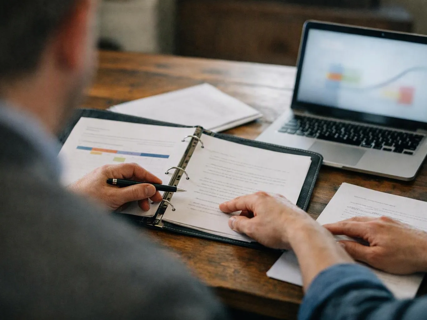 Two people review printed documents at a wooden table beside an open laptop and a binder