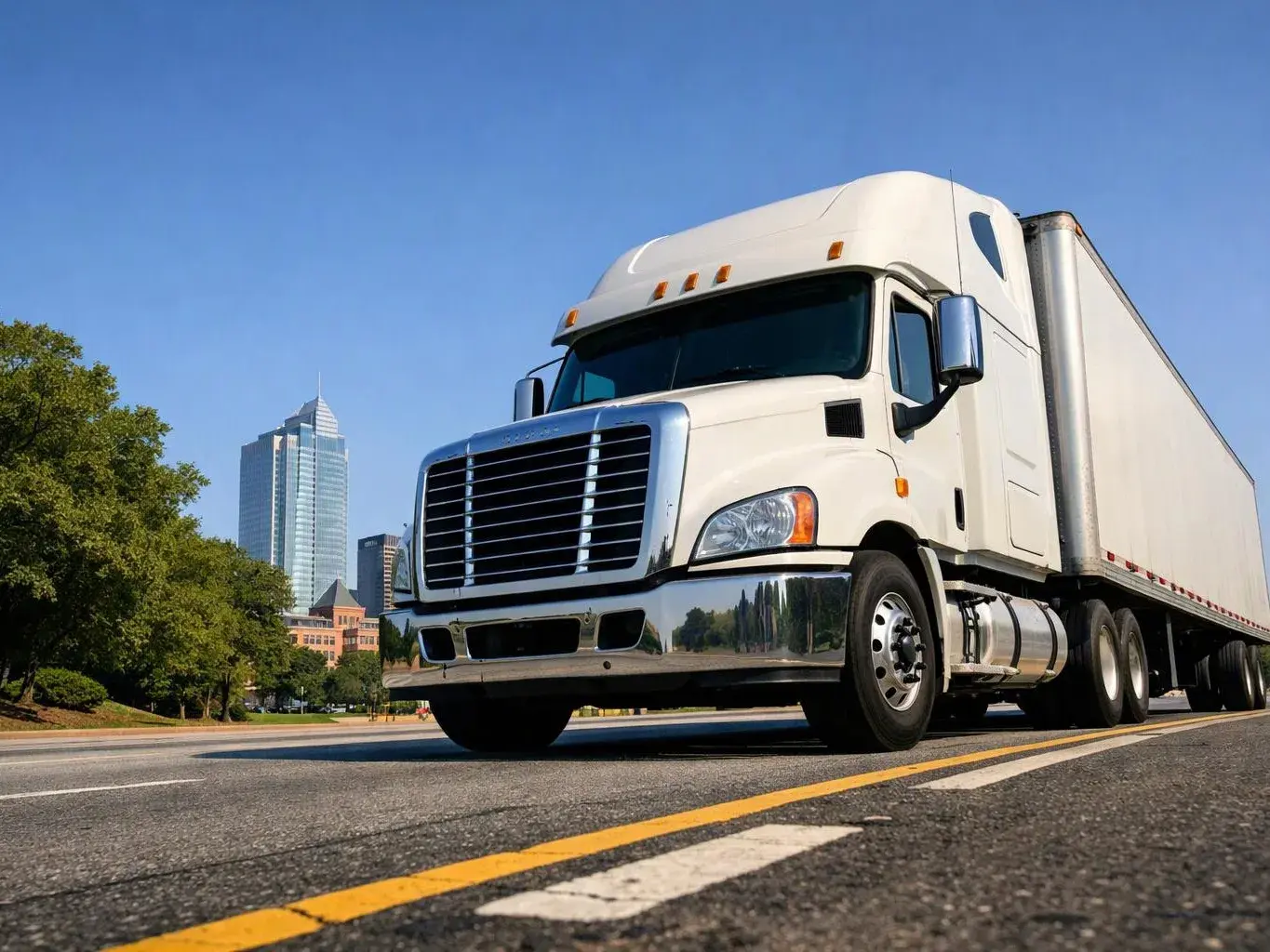 A white semi truck drives on a city street beside trees and tall buildings under a clear blue sky
